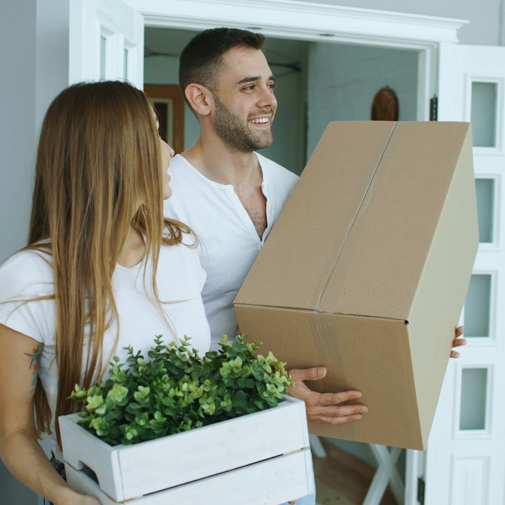 Two young adults smiling as they carry boxes and a plant into their new home